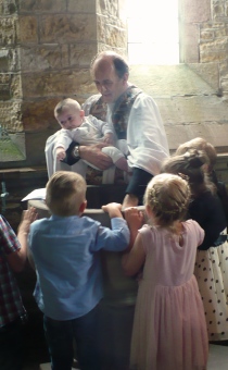 Priest baptising baby, children around font